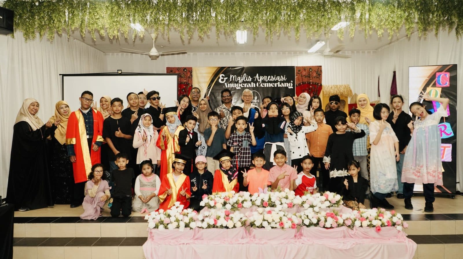 A large group photo of learners, families and teachers waving on stage