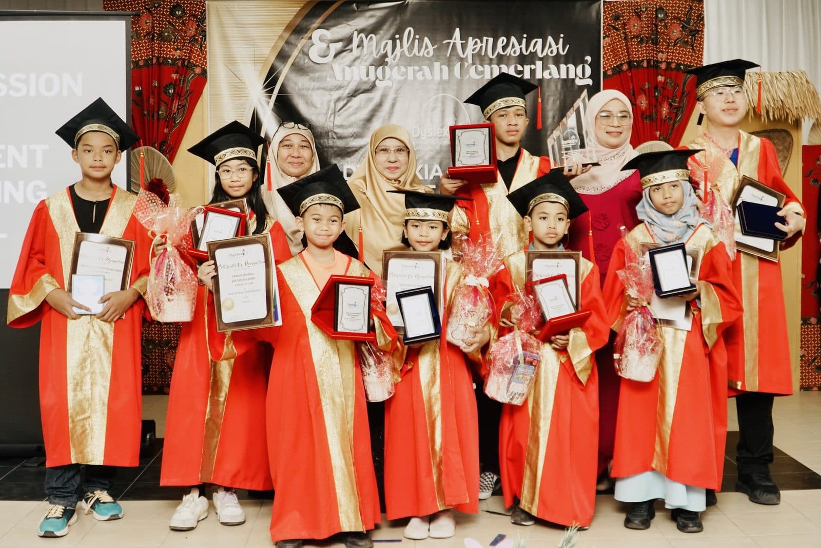 Graduates in red and gold gowns and caps holding their certificates on stage with their teachers