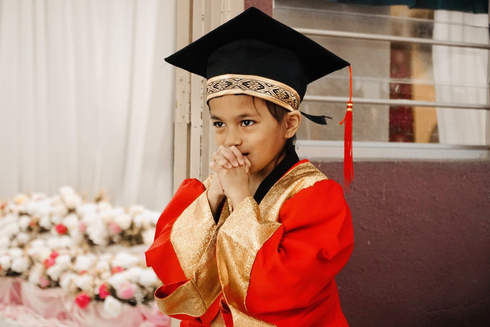 A young learner in a graduation gown and cap, hands clasped with excitement